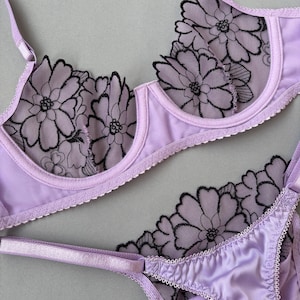 Puede incluir: Un conjunto de lencería color lavanda compuesto por un sujetador con aros y una tanga a juego. Ambas piezas presentan paneles de malla transparente con bordados florales negros, tirantes finos ajustables y bordes festoneados.