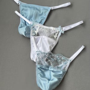 Könnte beinhalten: Drei String-Unterhosen in Blau- und Weißtönen. Die oberen und unteren Strings sind aus hellblauem Satin. Der mittlere String ist weiß mit Spitzendetails und kleinen blauen Schleifen. Alle haben weiße Träger.