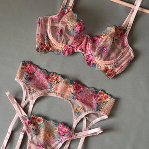 May include: A three-piece lingerie set in a soft pink hue, featuring intricate floral embroidery in shades of pink, orange, yellow, and teal. The set includes a bra, garter belt, and thong, all with delicate straps and scalloped edges.