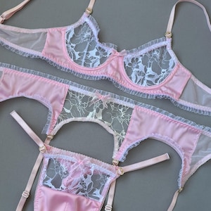 Può includere: Un completo di lingerie coordinato composto da reggiseno, reggicalze e perizoma rosa chiaro. Ogni capo è realizzato in raso rosa chiaro e pizzo floreale bianco, arricchito da delicati volant, piccoli fiocchi e dettagli dorati a forma di cuore.