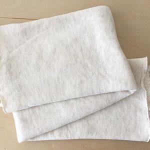 Puede incluir: Un trozo de tela de color blanco roto doblado con una textura ligeramente rugosa. La tela tiene un borde sin rematar.