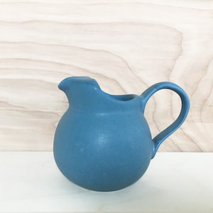 Può includere: Un creamer in ceramica blu con corpo arrotondato e manico. Il creamer è appoggiato su una superficie in legno chiaro.