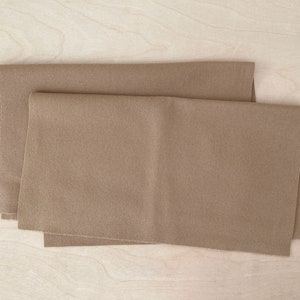 Puede incluir: Dos piezas de tela de fieltro marrón dobladas juntas. La tela es lisa y tiene un brillo sutil.