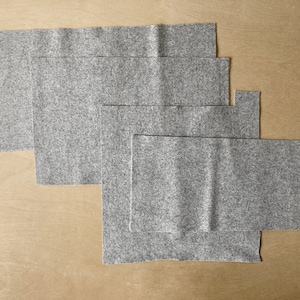 Puede incluir: Varios rectángulos de fieltro gris de diferentes tamaños se disponen sobre una superficie marrón claro. El fieltro tiene una textura moteada. La disposición sugiere un proyecto de manualidades, como costura.