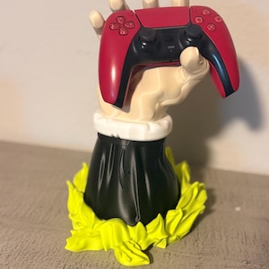 Puede incluir: Un mando de videojuegos rojo y negro es sostenido por una escultura de mano color crema. La mano descansa sobre una base negra con un cuello blanco y está rodeada por un diseño amarillo brillante en forma de hojas.