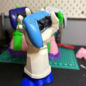 Puede incluir: Una mano blanca impresa en 3D sosteniendo un mando de Nintendo Switch azul. La mano está de pie sobre una base azul.