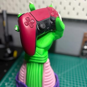 Può includere: Un supporto a forma di mano verde e rosa con una base viola tiene un controller per videogiochi rosso. Il supporto è progettato per assomigliare a un personaggio immaginario.