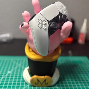 Può includere: Un controller per videogiochi bianco e nero è appoggiato su un supporto a forma di mano rosa. Il supporto ha una base nera con finiture dorate e un emblema bianco con un simbolo "M" dorato. Il controller ha un layout di pulsanti simmetrico.