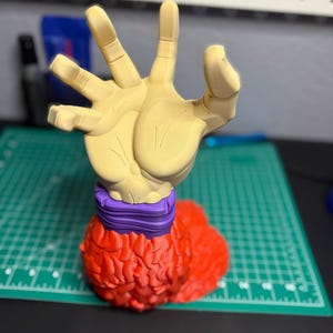 Puede incluir: Una figura impresa en 3D de una mano que se levanta, con una banda morada alrededor de la muñeca y una base roja texturizada.