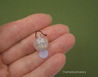 May include: A miniature clear plastic lantern with a brown string handle and a small, round, light purple base. The lantern has a silver-colored interior and is held in a hand against a green background. The Painted Canary is written in the bottom right corner.