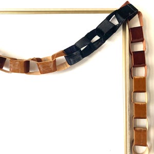 May include: A decorative chain made of connected velvet loops in shades of brown and black. The chain is hanging from a gold-colored rectangular frame, creating a unique visual display. The velvet material adds a touch of luxury.