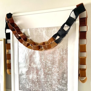 May include: A decorative chain made of velvet in shades of brown and black hangs in front of a framed artwork. The chain is draped over the top of the frame and hangs down on either side. The artwork has a silver and white textured background.
