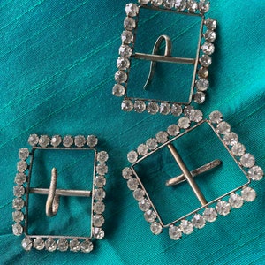 May include: Three silver-toned square buckles with a clear rhinestone border. The buckles have a simple, straight design.