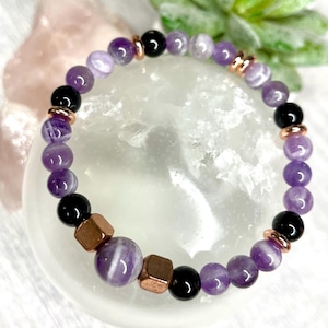 May include: A purple and white amethyst gemstone bracelet with black onyx beads and rose gold accents. The bracelet is on a white background.