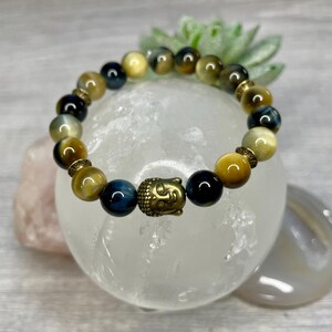 May include: A beaded bracelet featuring tiger's eye stones in shades of gold, blue, and black. The bracelet includes a gold-colored Buddha head charm and decorative spacers. The bracelet is displayed on a white, round surface.