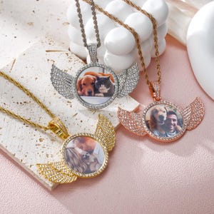 Puede incluir: Tres collares de oro, plata y oro rosa con un colgante de foto redondo que presenta una imagen de un perro y un gato, un perro y una persona, y una pareja con un perro pequeño. Cada colgante tiene un par de alas con piedras transparentes.