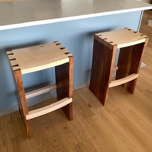 Peut inclure: Deux tabourets de bar en bois avec des sièges de couleur claire et des pieds marron foncé. Les pieds sont reliés par un repose-pieds incurvé de couleur claire. Les tabourets ont un design moderne et sont placés sur un plancher en bois.