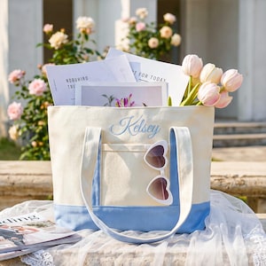 May include: A cream and light blue canvas tote bag featuring the embroidered name Kelsey in blue script. The bag has two shoulder straps, a front pocket, and a pair of white heart-shaped sunglasses hanging from the pocket edge. The bag is filled with papers and light pink tulips.