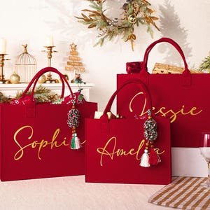 May include: Three red tote bags with gold script names: Sophie, Amelia, and Jessie. Each bag has a matching handle and a decorative tassel. The bags are displayed on a white surface with Christmas decorations in the background.