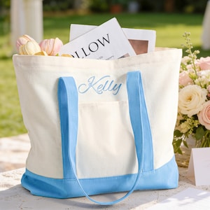 以下が含まれることがあります： クリーム色のキャンバス地のトートバッグ。水色のトリムとハンドル、水色の文字で「Kelly」と刺繍されています。バッグには花と読書用の資料が入っています。バッグの底は水色です。