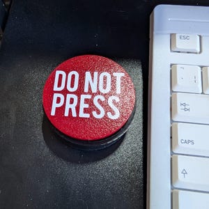 May include: A red circular button with the words "DO NOT PRESS" in white block letters. The button is on a black surface, with a white keyboard visible in the background.