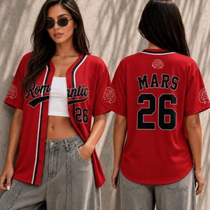 May include: A red baseball jersey featuring black and white trim, the text Romantic on the front, the number 26 on the front and back, and the text MARS on the back. The shirt includes white rose graphics on the sleeves and back neckline.