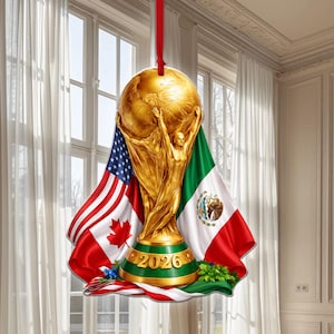 May include: A gold-colored ornament featuring the FIFA World Cup trophy, with the flags of the United States, Canada, and Mexico. The year "2026" is displayed on the base. A red ribbon is attached for hanging.