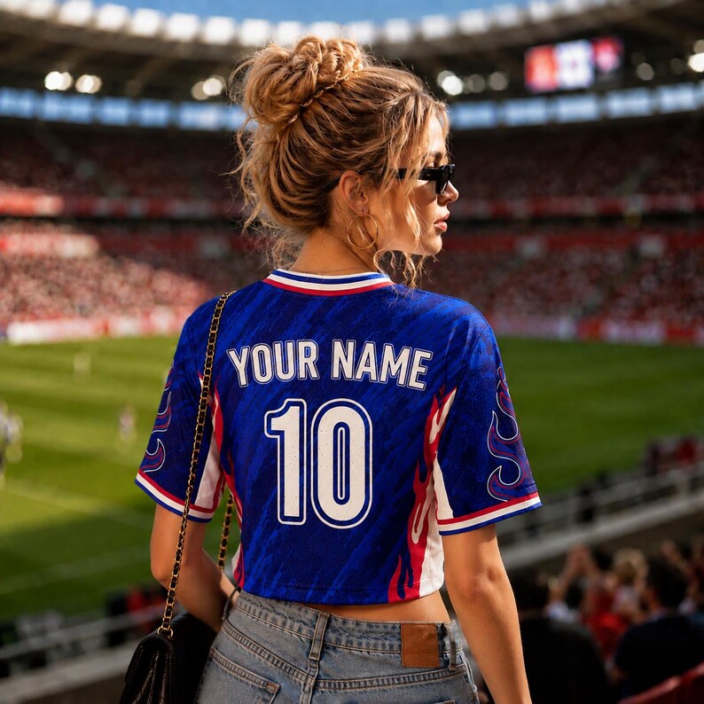 May include: A cropped blue jersey with the text "YOUR NAME" and the number "10" in white. The jersey has red and white accents and flame designs on the sleeves. The person is wearing blue jeans and sunglasses.
