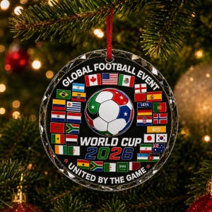 May include: A round, clear acrylic ornament featuring a central soccer ball graphic surrounded by numerous international flags. The text reads GLOBAL FOOTBALL EVENT, WORLD CUP 2026, and UNITED BY THE GAME. It hangs from a thin red string.