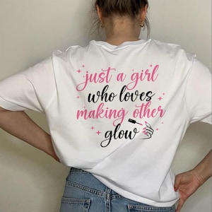 Puede incluir: Camiseta blanca con la frase "just a girl who loves making other glow" en letras rosas y negras. El diseño incluye estrellas rosas y una ilustración de un frasco de esmalte de uñas y una mano.