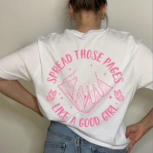 以下が含まれることがあります： 背中にピンク色のグラフィックが入った白いTシャツ。グラフィックは、手と「SPREAD THOSE PAGES LIKE A GOOD GIRL」というテキストが入った本が特徴です。2匹のピンク色の蝶と星がテキストを囲んでいます。