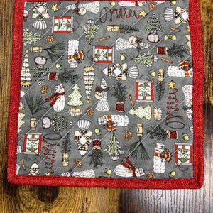 May include: Square quilted Christmas-themed potholder with a gray background and a red border. The fabric features various holiday illustrations, including snowmen, Christmas trees, and ornaments. The design is stitched with a diagonal pattern.