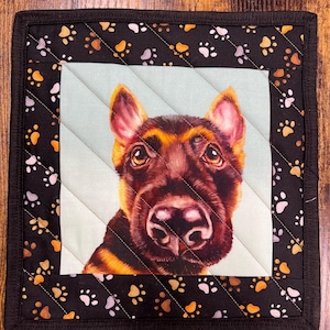 May include: Square quilted pot holder featuring a close-up of a German Shepherd dog's face. The dog has brown and black fur, with a light blue background. The border is black with paw prints in white, brown, and gold.