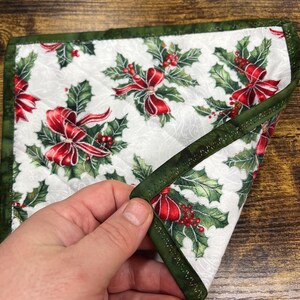 May include: A quilted Christmas-themed square featuring a white background with a pattern of red bows, holly leaves, and berries. The edges are trimmed with a dark green fabric. The item is being held by a hand.