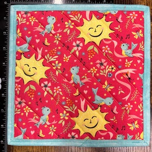 May include: Square quilted hot pad with a red background featuring smiling yellow suns, light blue birds, butterflies, and musical notes. The edges are trimmed with a light blue border. The hot pad measures approximately 8 inches square.