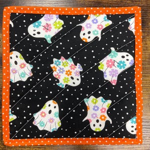 May include: Square quilted fabric coaster with a black background and white polka dots. The coaster features ghost shapes with floral designs in various colors. The border is orange with white polka dots.