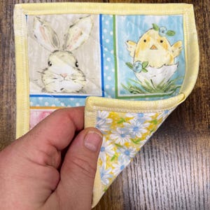 May include: A small, square quilted item with a yellow border. The front features a bunny and a chick in an egg on a blue background. The reverse side has a yellow floral pattern with blue flowers. The item is held by a hand.