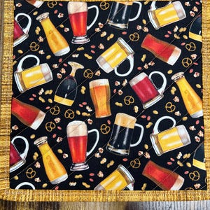 May include: A quilted square coaster with a black background featuring beer mugs, bottles, pretzels, and peanuts. The coaster has a gold border and is likely made of fabric. The design is a repeating pattern.