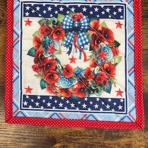 May include: A square quilted potholder featuring a central wreath of red poppies and blue hydrangeas with a blue and white gingham bow. The design includes white stars on a dark blue border, a light blue plaid frame, and a red polka dot binding.