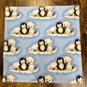 May include: Square, light blue fabric with a repeating pattern of polar bears and penguins on clouds. The polar bears are white, and the penguins are black and white. The fabric has a white stitched border.