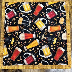 May include: A square quilted fabric coaster with a black background featuring a pattern of beer mugs, pretzels, and peanuts. The mugs are filled with various types of beer. The border is a gold color.