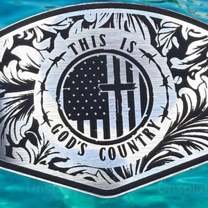 May include: Silver-colored belt buckle with a decorative floral border. The center features the American flag with a cross, surrounded by barbed wire and the text "THIS IS GOD'S COUNTRY."