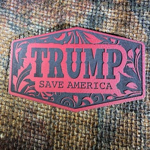 May include: A red and black decorative plaque with the word "TRUMP" in bold letters and the phrase "SAVE AMERICA" below. The plaque has a decorative border with floral accents.