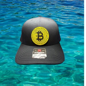 Puede incluir: Gorra de camionero negra con un parche con el logotipo de Bitcoin amarillo. La gorra tiene una visera curva y una parte trasera de malla. La etiqueta de la marca Richardson es visible. La gorra está sobre un fondo de agua azul.