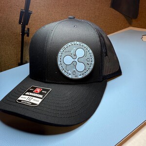 Puede incluir: Gorra de camionero negra Richardson 112 con parte trasera de malla y visera curva. La gorra presenta un parche circular con un símbolo de criptomoneda Ripple azul claro y texto. La gorra es ajustable.