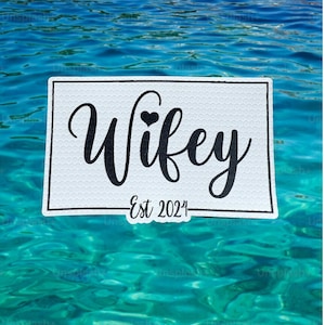 May include: A white sticker with the word "Wifey" in black script, a small heart, and "Est 2024". The sticker has a textured background and is set against a backdrop of blue water.