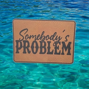 May include: A rectangular brown sign with the words "Somebody's PROBLEM" in black script. The sign is set against a backdrop of blue water.
