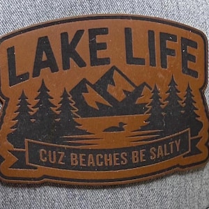 May include: A brown leather patch with the words "LAKE LIFE" above a mountain and lake scene with pine trees and a loon. The banner below reads "CUZ BEACHES BE SALTY". The patch is on a grey fabric background.