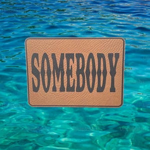 May include: A rectangular brown leather patch with the word "SOMEBODY" in bold, black letters. The patch is set against a backdrop of clear, turquoise water.