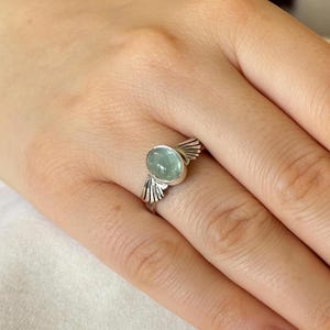 Op de afbeelding: Een zilveren ring met een lichtblauwe ovale edelsteen en vleugelvormige accenten.